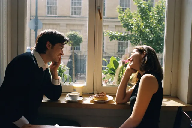 Two people lean in and laugh across a café window counter in the morning light.