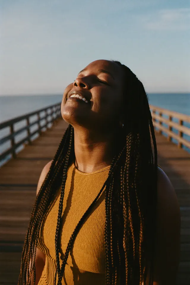 A young woman with long braids tips her face to the sun, eyes closed and smiling, on a pier by the sea.